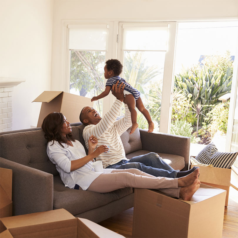 A mother and father sit on a couch lifting up their child and laughing, surrounded by open moving boxes.