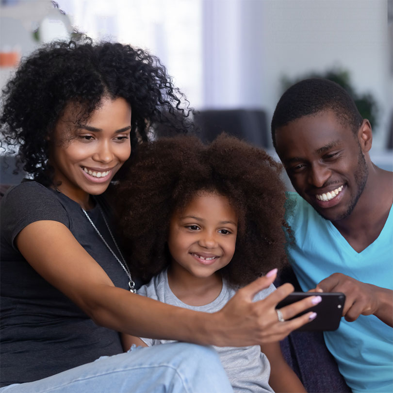 A mother, father and young daughter huddle around a smartphone, smiling.