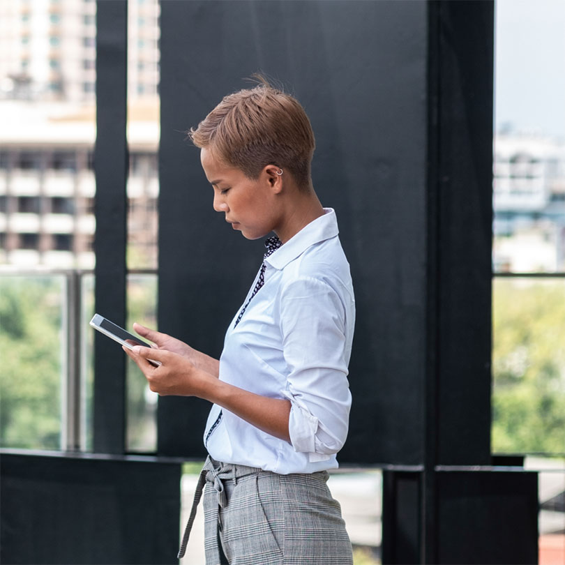 A woman in business attire stands outside looking down at her phone.