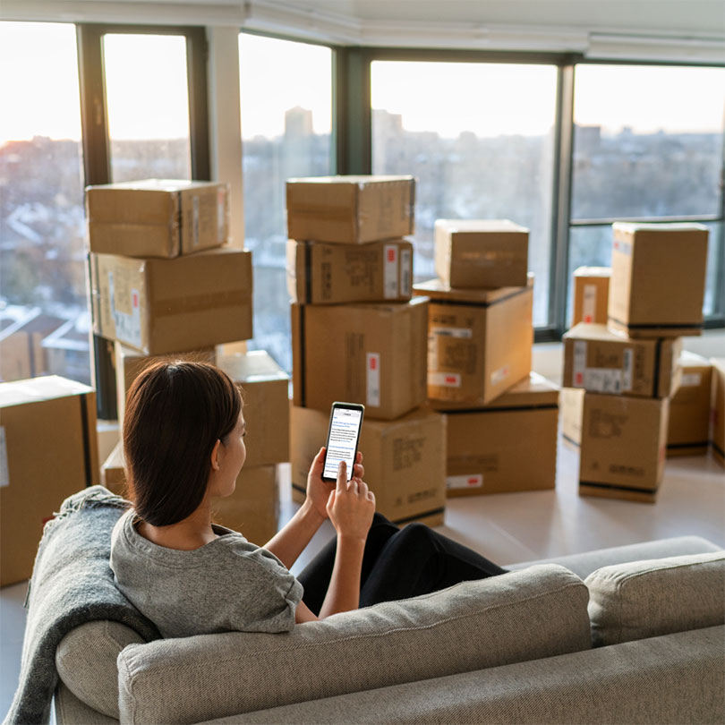 A woman sits on a couch looking at her phone, surrounded by stacks of moving boxes.